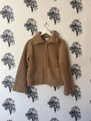 Brun/beige teddyjacka med dragkedja - Mysig brun/beige teddyjacka med hög krage och dragkedja framtill. Jackan har två sidofickor och är tillverkad i ett fluffigt, mjukt material som ger en avslappnad och trendig look. Perfekt för kyliga dagar och enkel att matcha med olika outfits.