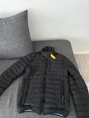 Svart pufferjacka från Parajumper  - Snygg svart pufferjacka från Parajumper  med quiltad design och hög krage. Jackan har dragkedja framtill, två sidofickor med dragkedja och en bröstficka. Insidan är ljusgrå och materialet är lätt och värmande, perfekt för kalla dagar.