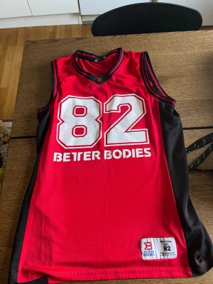 Röd basketlinne Better Bodies - Snyggt rött basketlinne från Better Bodies med svarta sidopaneler och vita detaljer. Stort tryck med siffran 82 och texten 'Better Bodies' på framsidan. Tillverkat i meshmaterial som andas, perfekt för träning eller sport.