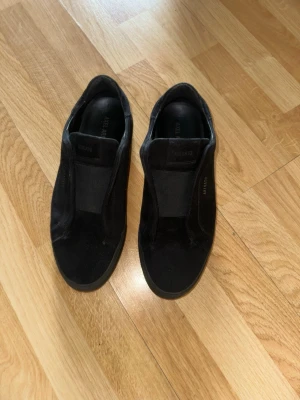 Svarta slip-on sneakers från Axel Arigato - Stilrena svarta slip-on sneakers från Axel Arigato i mocka. Skorna har en minimalistisk design utan snörning och en tjock sula som ger en modern look. Perfekta för dig som gillar enkel och clean stil.