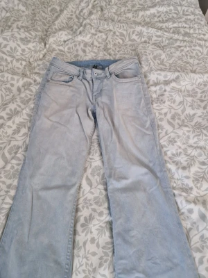 Ljusblå bootcut jeans - Säljer ett par ljusblå bootcut jeans i klassisk femficksmodell med knappstängning och bälteshällor. Jeansen har bakfickor med lock och knapp, vilket ger en unik touch. Tillverkade i mjuk denim som är skön att bära.