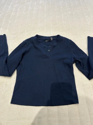 Mörkblå ribbad topp från ASOS DESIGN - Mörkblå långärmad topp från ASOS DESIGN i ribbad bomull med två knappar vid halsen. Enkel och stilren modell som passar perfekt till jeans eller kjol. Skön och mjuk kvalitet med normal passform.