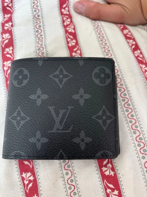  svart monogram plånbok - Snygg svart plånbok med klassiskt monogrammönster i grått. Plånboken har flera kortfack, ett fönster för ID och är tillverkad i tåligt läder. Perfekt för dig som vill ha en stilren och lyxig accessoar.