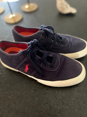 Marinblå sneakers från Polo Ralph Lauren - Snygga marinblå sneakers från Polo Ralph Lauren med klassisk låg profil och vit sula. Skorna har mörkblå snören, röd insida och den ikoniska röda Polo-loggan broderad på sidan. Tillverkade i canvas för en avslappnad och stilren look. #sneaker #poloralphlauren