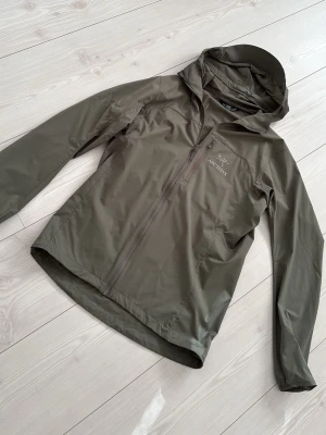  Arc'teryx vindjacka  - Snygg olivgrön vindjacka från Arc'teryx med hel dragkedja framtill, huva och diskret logga på bröstet. Tillverkad i lätt och slitstarkt material som står emot vind. Perfekt för dig som vill ha en stilren och funktionell jacka.