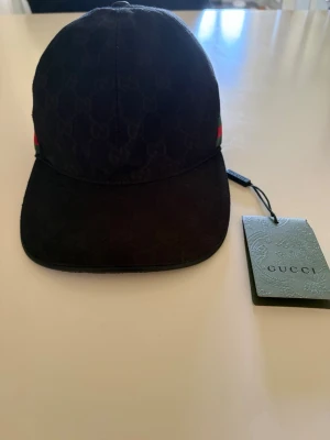 Svart keps från Gucci med logga - Snygg svart keps från Gucci med diskret GG-monogram över hela tyget och klassiska gröna och röda ränder på sidan. Kepsen har böjd skärm och justerbar passform. Tillverkad i slitstark bomullscanvas och levereras med originaltagg. Kepsen är helt ny och pris kan även diskuteras.