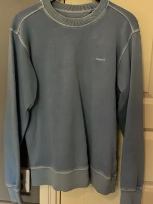 Ljusblå sweatshirt från GANT - Snygg ljusblå sweatshirt från GANT med rund halsringning och diskret logga på bröstet. Tröjan har ribbade muddar vid ärmslut, hals och nederkant. Perfekt för en avslappnad och stilren look.