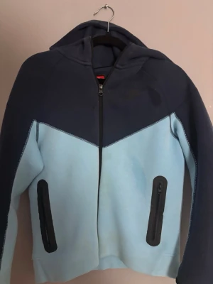 Nike blå hoodie med dragkedja - Snygg hoodie från Nike i två nyanser av blått. Modellen har hel dragkedja framtill, två svarta dragkedjeförsedda fickor och en stor huva. Tillverkad i mjukt material som känns skönt mot huden. Perfekt för en avslappnad streetwear-look.     Det som är lite mörkare på två delar är bara vatten 