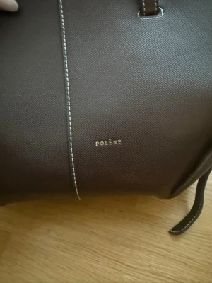 Mörkbrun handväska från Polène - Snygg mörkbrun handväska från Polène i läder med vit kontrastsöm. Väskan har två handtag och en stilren, strukturerad form. Perfekt för dig som gillar minimalistisk design och vill ha en väska som sticker ut med sina detaljer.                                                   I den mindre Cyme modellen, EJ ÄKTA 
