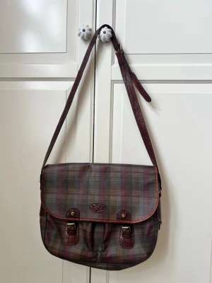     Äkta Mulberry handväska – Made in England - Säljer en Äkta klassisk handväska från Mulberry i rutigt tyg med bruna läderdetaljer och spännen framtill. Väskan har justerbar axelrem och präglad Mulberry-logga på insidan. Perfekt för dig som gillar vintage och brittisk stil. Ser ut som ny Se skick på bilderna