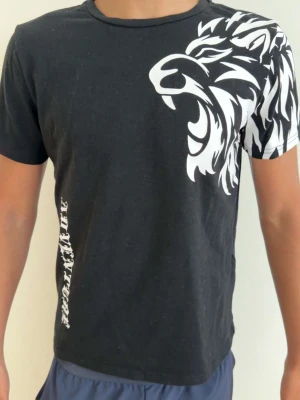 Svart t-shirt med lejontryck - Cool svart t-shirt med stort vitt lejontryck över axeln och bröstet. T-shirten har korta ärmar och rund halsringning. Snygg detalj med vit text längs sidan. Perfekt för dig som vill sticka ut med en unik design.