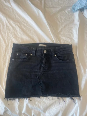 Svart jeanskjol  - Säljer en svart jeanskjol från gina med rå kant nertill och klassisk. Kjolen är kort och har en mörk tvätt, perfekt till sommaren. Passar dig som gillar en edgy och avslappnad stil.