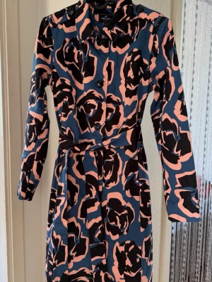 Mönstrad trenchcoat från Park Lane - Snygg trenchcoat från Park Lane med unikt abstrakt mönster i blått, rosa och svart. Jackan har knappar framtill, klassisk krage, axelklaffar och ett matchande knytbälte i midjan. Perfekt för dig som vill sticka ut med din stil.