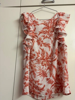 Vit blus med blommönster - Supersöt blus i vitt med blommönster och volangdetaljer längs axlarna. Blusen har en rak passform och är ärmlös, vilket gör den luftig och perfekt till sommaren. Materialet känns lätt och mjukt mot huden. Perfekt för dig som gillar romantiska detaljer!