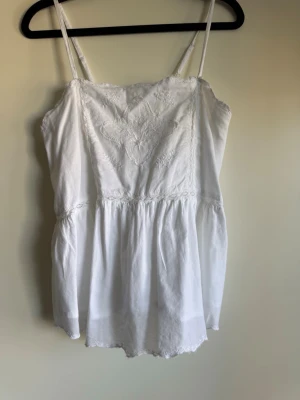 Vit broderad linnetopp med spets - Söt vit linnetopp med tunna axelband och broderade detaljer framtill. Toppen har spetskant och volang nertill för en romantisk känsla. Perfekt till sommaren och enkel att matcha med jeans eller kjol.