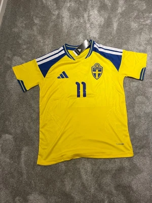 Svensk landslagströja Adidas nr 11 - Gul och blå svensk landslagströja från Adidas med nummer 11 och namnet Larsson på ryggen. Tröjan har korta ärmar, broderad landslagssköld och klassiska Adidas-ränder på axlarna. Tillverkad i lätt och ventilerande polyester, perfekt för fotboll.