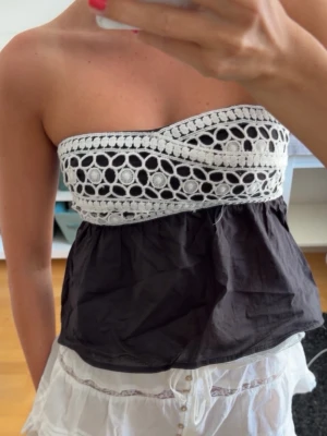 Svart och vit tubtopp - Säljer en snygg axelbandslös tubtopp i svart och vitt. Överdelen har ett virkat, spetsliknande mönster med cirklar och underdelen är svart och luftig. Perfekt för sommardagar och enkel att matcha med kjol eller jeans.