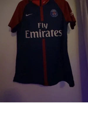 PSG Neymar Jr matchtröja blå/röd - Snygg PSG matchtröja från Nike i blått och rött med Neymar Jr och nummer 10 på ryggen. Tröjan har Fly Emirates-tryck på framsidan och klubbmärke på bröstet. Perfekt för fotbollsfans som vill visa sitt stöd för Paris Saint-Germain.
