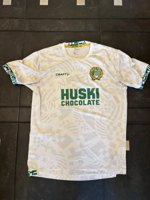 Vit Hammarby matchtröja från Craft - Säljer en vit Hammarby fotbollströja från Craft med gröna detaljer och klubbmärke på bröstet. Tryck med 'HUSKI CHOCOLATE' på framsidan och mönstrade muddar i grönt och vitt. Texten 'UNDER GRÖNVIT FLAGG' finns på insidan av kragen.