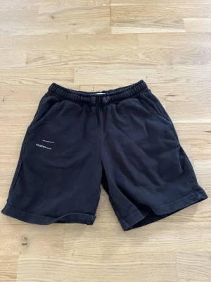 Svarta shorts med resårmidja - Svarta shorts med avslappnad passform och elastisk midja. Två sidofickor och diskret tryck på vänster ben. Perfekta för sommardagar eller chill hemma. Tillverkade i mjuk bomull för maximal komfort.