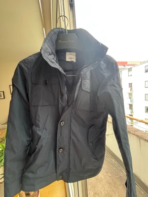 Mörkblå vindjacka från J.Lindeberg - Stilren mörkblå vindjacka från J.Lindeberg med hög krage, bröstfickor med knappar och dragkedja framtill. Jackan har en klassisk passform och är tillverkad i ett lätt syntetmaterial, perfekt för vår och höst. Pris kan diskuteras!