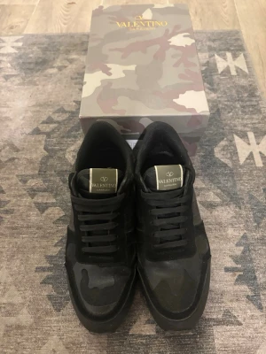 Valentino Garavani rockrunners - Snygga svarta sneakers från Valentino med camo-mönster och detaljer i mocka och läder. Skorna har klassisk låg profil, svarta skosnören och coola nitar på hälen. Perfekta för dig som vill ha en exklusiv och trendig look.