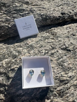 Caroline Svedbom örhängen blå kristall - Snygga örhängen från Caroline Svedbom med droppformade, ljusblå kristaller infattade i guldfärgad metall. Perfekta för att ge din look lite extra bling och färg. Kommer i originalask.