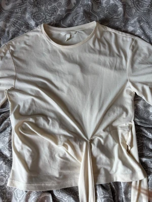 Krämvit topp med knyt från H&M - Säljer en krämvit topp från H&M med rund halsringning och snygg knyt-detalj i midjan. Toppen har korta ärmar och är gjord i mjuk bomull, perfekt för en clean och enkel look. Denna tröjan är använd fåtal gånger. Skriv vid frågor!!🤍