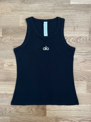 Svart ribbad tanktop från Alo - Snygg svart ribbad tanktop från Alo med rund halsringning och brottarrygg. Toppen har en tight passform och är tillverkad i mjuk bomullsmix. Perfekt för en sportig och trendig look.