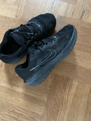 Svarta Nike sneakers med mesh - Snygga helsvarta Nike sneakers med klassisk swoosh på sidan. Ovandelen är i mesh och syntet för bra andningsförmåga och komfort. Skorna har svarta snören och en robust sula som passar perfekt till streetwear och vardagsstil.