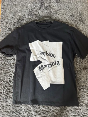 Maison Margiela svart t-shirt - Säljer en svart t-shirt från Maison Margiela med stor vit grafisk print framtill där märkesnamnet syns. T-shirten har klassisk passform, rund hals och korta ärmar. Tillverkad i mjuk bomull som känns skön mot huden. Storlek L (48) men passar för XL. Pris kan diskuteras.