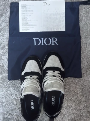 Dior B25 - Snygga Dior B25 sneakers i svart och vitt med mesh och mockadetaljer. Skorna har tjock vit sula, vita skosnören och Dior-logga på plösen och innersulan. Perfekta för dig som vill ha en exklusiv och trendig look. Tyvärr ingen box kvar, bara påse och kuvert