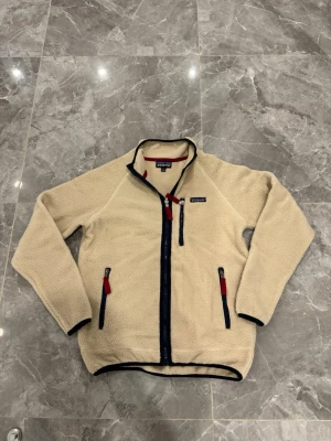 Beige fleecejacka från Patagonia - Mysig beige fleecejacka från Patagonia med marinblå detaljer och röda dragkedjor. Jackan har hög krage, tre fickor med dragkedja och är tillverkad i mjukt fleecematerial. Perfekt för lager-på-lager och outdoor-vibes.