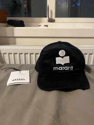 Isabel Marant keps - Snygg svart keps i manchestertyg från Marant med vit broderad logga och text framtill. Klassisk böjd skärm och justerbar baksida. Perfekt accessoar för en trendig och avslappnad stil.