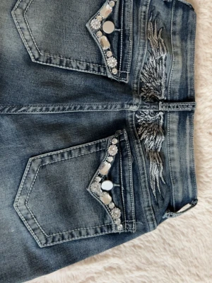 Blå bootcut jeans med strassdetaljer - Snygga blå bootcut jeans med broderade vingar på bakfickorna och coola strassdetaljer samt silvriga knappar. 