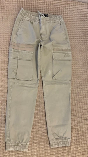 Beige cargobyxor med resår från Pull & Bear - Snygga beige cargobyxor med resår i midja och benslut. Byxorna har flera stora fickor på sidorna och bak, samt detaljerade sömmar. Perfekta för en avslappnad streetwear-look. Tillverkade i ett mjukt bomullsmaterial.