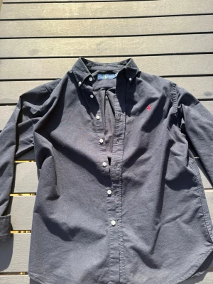 Ralph Lauren skjorta  - Klassisk mörkblå skjorta från Ralph Lauren med knappkrage och vit knappstängning. Skjortan har lång ärm och den ikoniska röda logotypen broderad på bröstet. Perfekt till jeans eller chinos för en clean look.