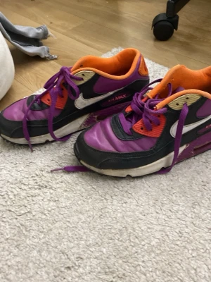 Nike Air Max lila och orange sneakers - Nike Air Max sneakers i lila, svart och orange med vita detaljer och lila skosnören. Klassisk Air Max-sula för extra komfort och retrovibe. Ovandelen är i syntetmaterial och mesh, med tydlig Nike-logga på sidan och AIR-detalj bak.