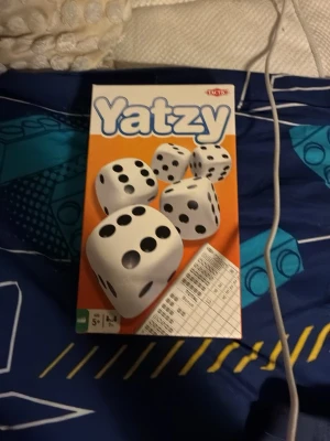 Yatzy sällskapsspel från Tactic - Klassiskt Yatzy sällskapsspel från Tactic. Innehåller fem vita tärningar med svarta prickar och ett poängblock. Förpackningen är orange och vit med stora tärningsbilder på framsidan. Perfekt för spelkvällar med vänner eller familj.