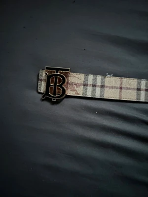 Beige rutigt Burberry bälte - Snyggt bälte från Burberry med klassiskt beige rutigt mönster och en stor metallspänne med Burberrys ikoniska TB-logga. Perfekt accessoar för att lyfta vilken outfit som helst.