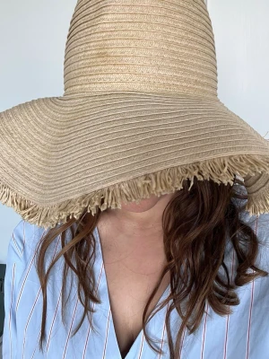 Straw hat from H&M - Säljer en stor, beige solhatt i flätad strå med bred brätte och fransad kant. Perfekt för soliga dagar och ger riktigt skön strandvibe. Hatten har en klassisk rund form och är lätt att matcha med somriga outfits.