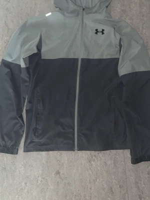 Grå vindjacka från Under Armour - Snygg tvåfärgad vindjacka från Under Armour i ljusgrått och mörkgrått. Jackan har huva, hel dragkedja framtill, två sidofickor och logga på bröstet. Tillverkad i lätt och vindtätt material, perfekt för blåsiga dagar.