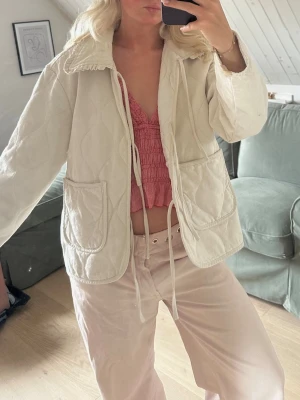 Beige quiltad jacka med snörning - Quiltad beige jacka med snörning framtill och två stora fickor. Jackan har en avslappnad passform och är tillverkad i ett mjukt material som känns skönt mot huden. Perfekt för lager på lager och ger en trendig look.