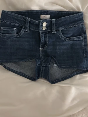 Mörkblå jeansshorts med dubbla knappar - Snygga mörkblå jeansshorts från Only med dubbla vita knappar framtill och klassiska fickor. Shortsen har dekorativa sömmar på bakfickorna och är i en tight passform. Perfekta för sommardagar och enkla att matcha med olika toppar.