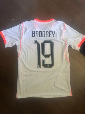 Holland fotbollströja Nike Brobbey - Säljer en vit Holland fotbollströja från Nike med Brobbey och nummer 19 på ryggen. Tröjan har orange detaljer, korta ärmar och är tillverkad i lätt och ventilerande material (AeroSwift). Perfekt för match eller träning.