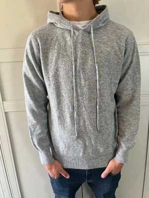 Ljusgrå hoodie i mjukt material - Säljer en stilren ljusgrå hoodie med huva och dragsnören. Tröjan är tillverkad i ett mjukt och skönt tyg, har ribbade muddar vid ärmslut och nederkant. Perfekt för en avslappnad look och passar till jeans eller joggers.