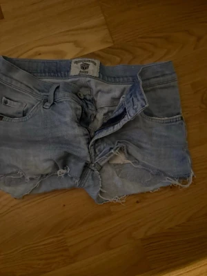 Ljusblå jeansshorts med råa kanter - Säljer ett par ljusblå jeansshorts från Cheap Monday. Shortsen har råa, fransiga kanter och klassisk femficksdesign. De är midwaist och har dragkedja framtill. Perfekta för sommaren och ger en avslappnad look.
