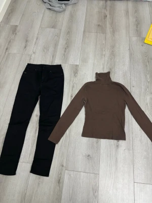Svarta raka jeans med fem fickor & Brun tröja med hög hals  - Säljer ett par svarta skinny jeans med klassisk rak passform och fem fickor. Jeansen har bälteshällor och knappgylf. Perfekta till en enkel vardagslook och passar till det mesta. Materialet känns som bomull med lite stretch. Jag säljer även en brun tröja med byxorna i material av bomull. Byxorna har två lite synliga fläckar på baksidan och tröjan har två hål vid nacken som går att sy ihop.