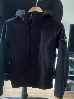 Svart vindjacka från Stone Island - Snygg svart vindjacka från Stone Island med huva och dragkedja framtill. Jackan har den klassiska logotypen på vänster ärm och en minimalistisk design. Perfekt för dig som vill ha en stilren och funktionell jacka till vardags.                                                                  Storlek - Junior 142/10 år,                                  Denna jackmodell från Stone Island tillverkad ej längre,                                                Priset på varan kan diskuteras.