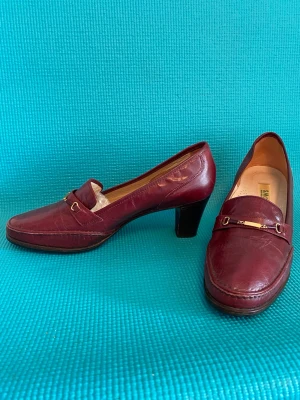 Vinröda loafers med klack i skinn 40,5 storlek - Snygga vinröda loafers från Santa Maria i äkta läder, tillverkade i Italien. Skorna har en klassisk penny loafer-design med guldfärgad metall-detalj och en stadig klack. Perfekta för dig som vill ha en stilren och tidlös look. Innermått ca 26 cm, klacken 4,6 cm.